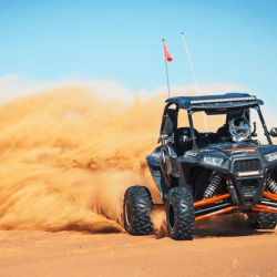 A person driving a dune buggy through the rolling sand dunes of the Dubai desert, with clouds of sand in the air and the desert landscape in the background