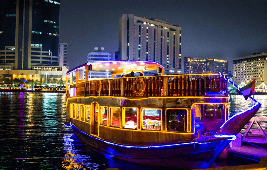 A traditional dhow boat lit up with colorful lights and decorations, sailing down the Dubai Water Canal with the city's modern skyline in the background